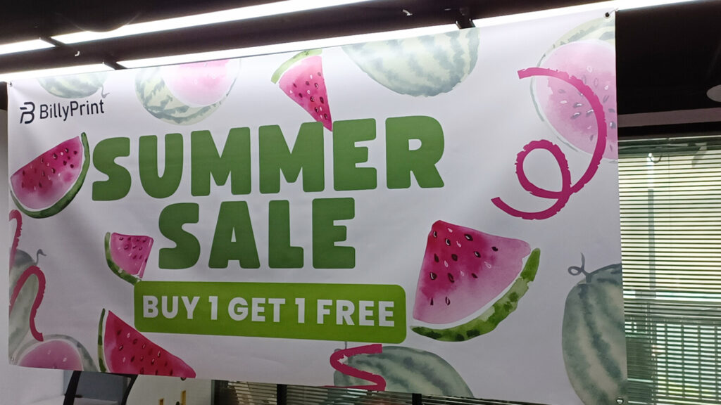 Close-up view of a green and pink double-sided vinyl banner by BillyPrint with a 'SUMMER SALE, BUY 1 GET 1 FREE' offer, displaying lime and watermelon graphics.